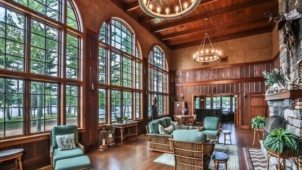 A spacious room with large windows, wooden floors, wicker furniture, and a fireplace. - Lakefront, Historic Estate on the Eagle River Chain of Lakes! Sleeps 42! (Eagle River)