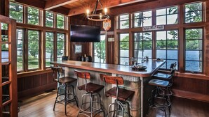 A rustic bar area with wooden walls, large windows overlooking a lake, a flat-screen TV, and metal bar stools. - Lakefront, Historic Estate on the Eagle River Chain of Lakes! Sleeps 42! (Eagle River)