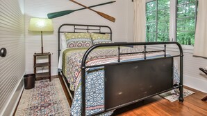 Un dormitorio con estructura de cama metálica, ropa de cama estampada, piso de madera, una mesita de noche, una lámpara y una ventana con vistas a zonas verdes.