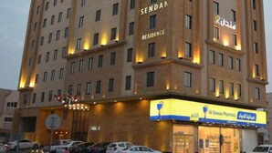 Un hotel de varios pisos con un llamativo letrero que dice 'SENDAH RESIDENCE', una farmacia y varios autos estacionados frente.