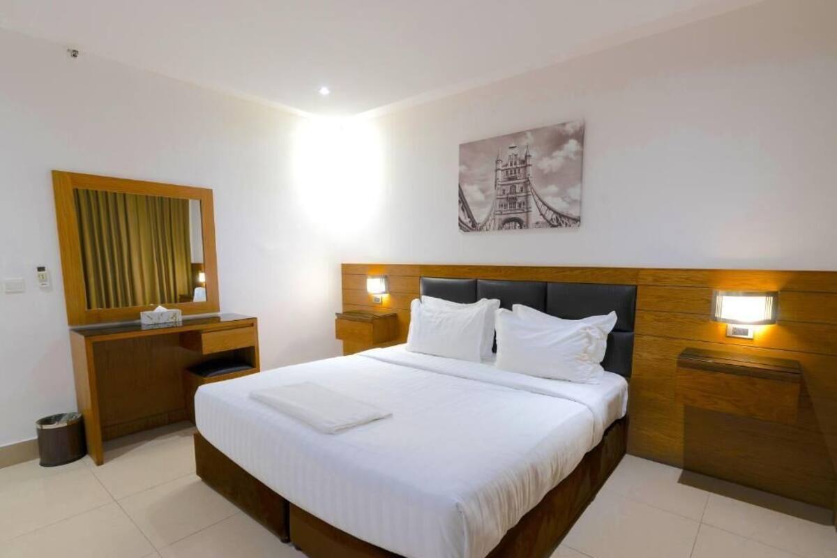 A neatly arranged hotel room with a large bed, bedside lamps, a wooden headboard, a mirror, and a wall-mounted picture.