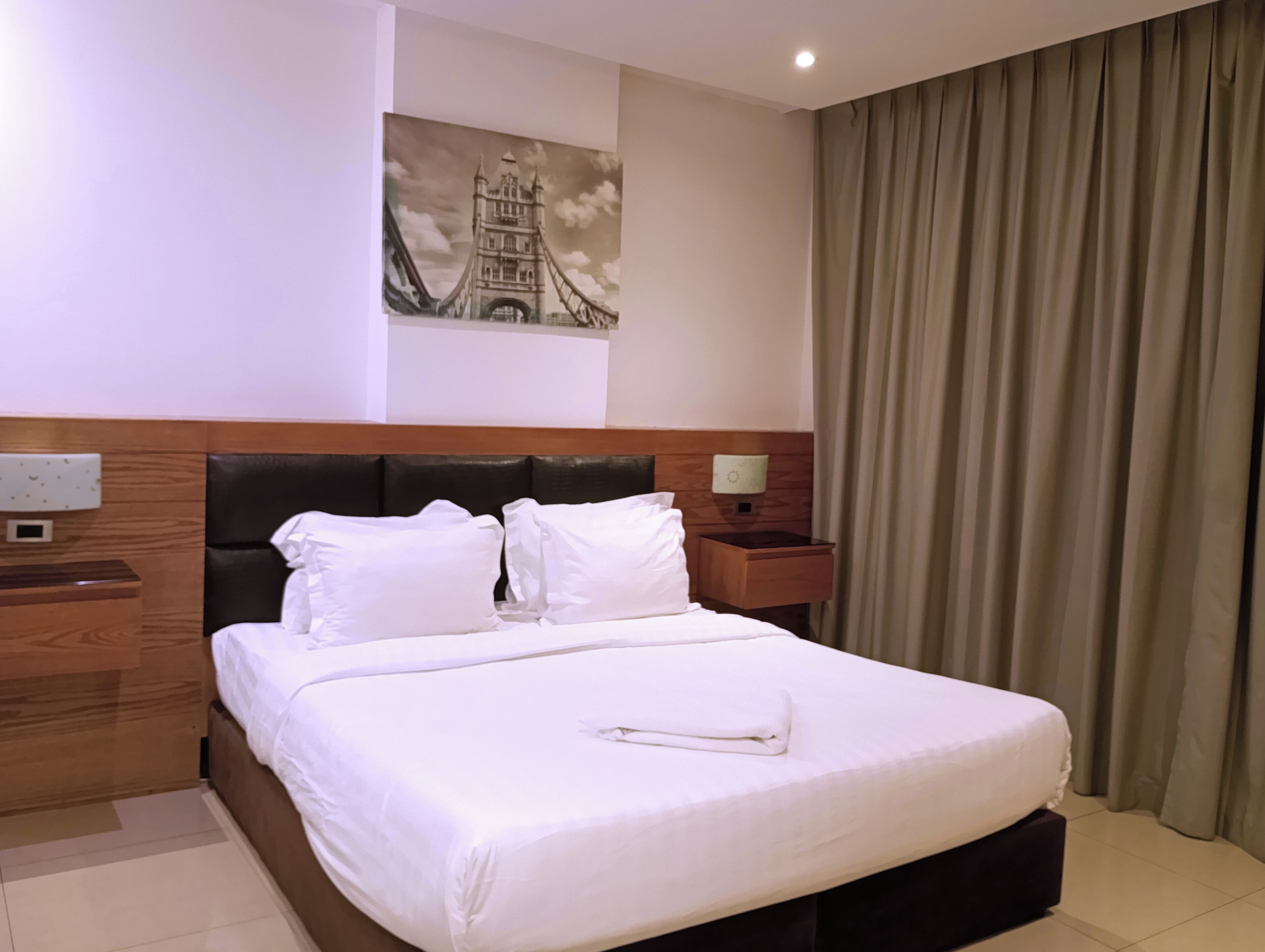 A hotel room with a bed, a nightstand, a lamp, a painting of a bridge, and a curtain.