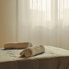 Un letto rifatto con due asciugamani arrotolati e tende leggere con una griglia decorativa alla finestra.