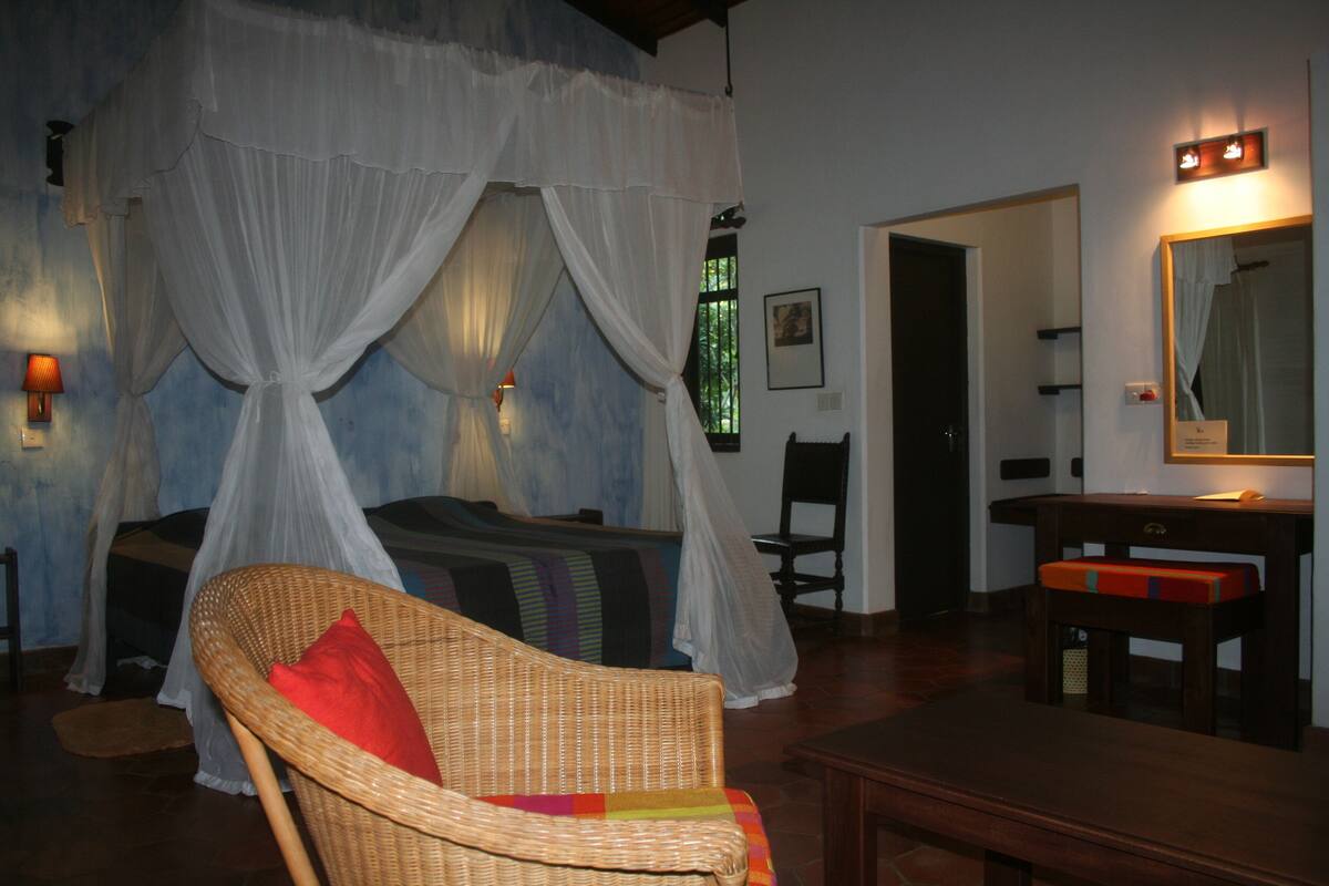 A room with a bed, a wicker chair with a red pillow, a wooden table, and a mirror on the wall.