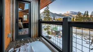 Una cabaña de madera con puerta de vidrio que da a un dormitorio, un balcón con barandilla de metal y vistas a una montaña nevada.