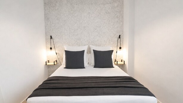 Een net opgemaakt bed met een witte matras en donkere beddengoed, met aan beide kanten twee bedlampjes.