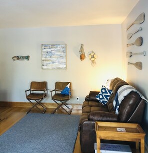 Uma sala de estar com um sofá de couro marrom, duas poltronas de couro marrom, um tapete azul e uma mesa de centro de madeira.