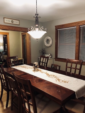 Uma sala de jantar com uma mesa de madeira, cadeiras, um candelabro, uma coroa de louros e uma janela com persianas.