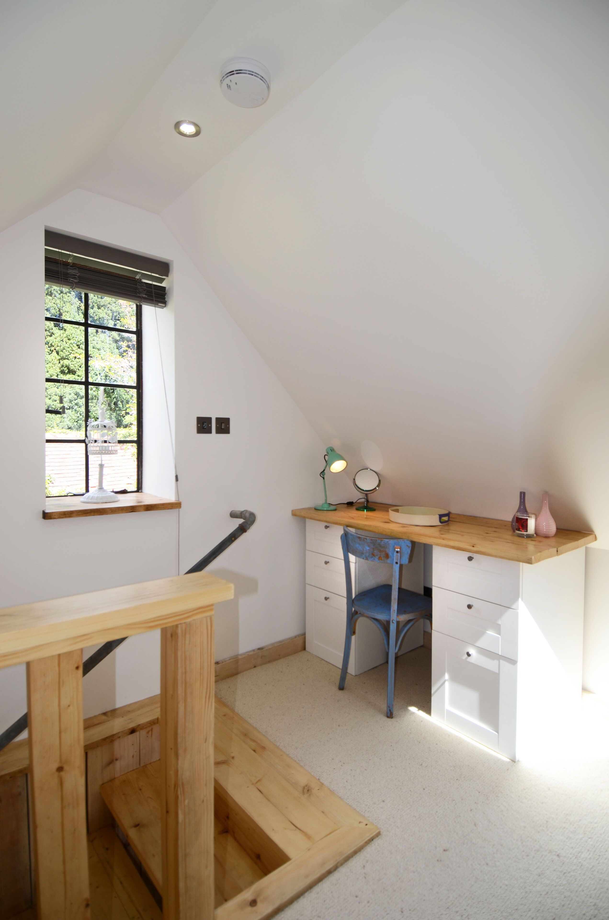 A small, well-lit room with a sloped ceiling, a wooden desk, a chair, a window with blinds, and a radiator.