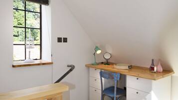 A small, well-lit room with a sloped ceiling, a wooden desk, a chair, a window with blinds, and a radiator.