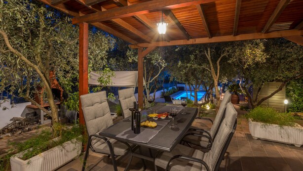 An outdoor patio with a table set for dining, surrounded by chairs, and a pool in the background. - Detached House on a plot of 500m2 with private pool and a walled Oliva garden (Stari Grad)