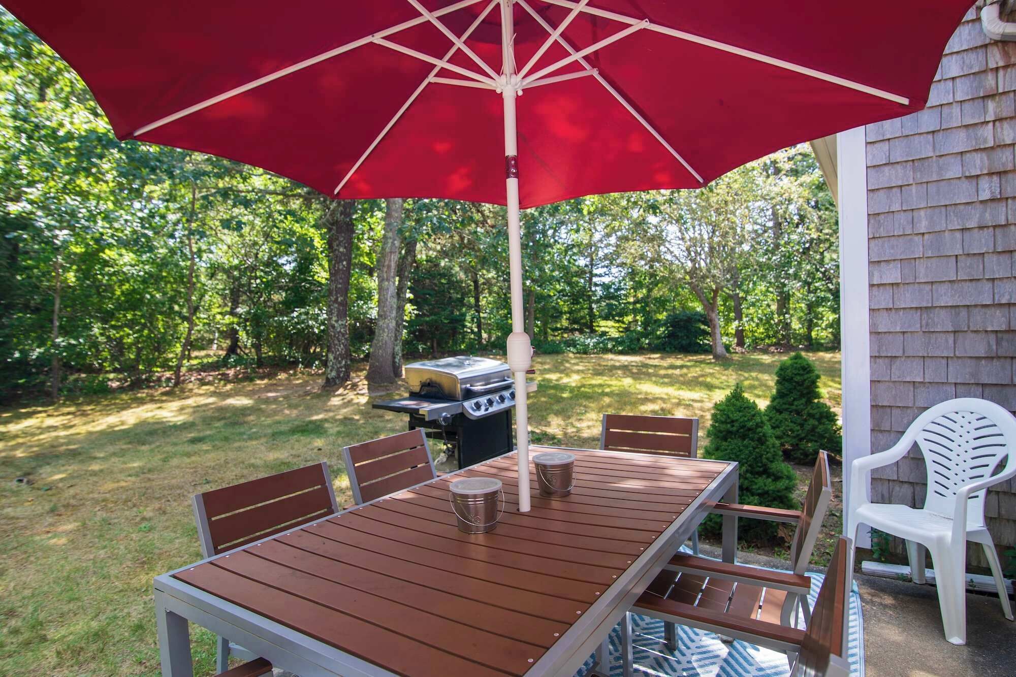 Une table de patio en bois, quatre chaises, un parasol rouge et un barbecue dans un jardin.
