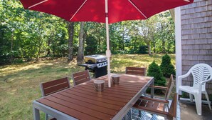 Une table de patio en bois, quatre chaises, un parasol rouge et un barbecue dans un jardin.