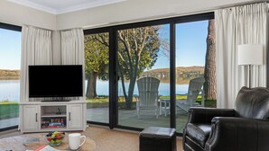 Een woonkamer met een flatscreen televisie, een bijzettafeltje met een schaal fruit, een tijdschrift en afstandsbedieningen. Er is een leren bank en een grote schuifglazen deur die leidt naar een buitenruimte met stoelen en uitzicht op een meer.