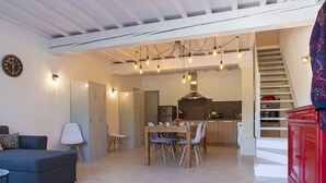 Un grand espace de vie comprenant un canapé, une table à manger et des chaises. On y trouve également une cuisine équipée d’une cuisinière et de placards. Un escalier est visible au fond.