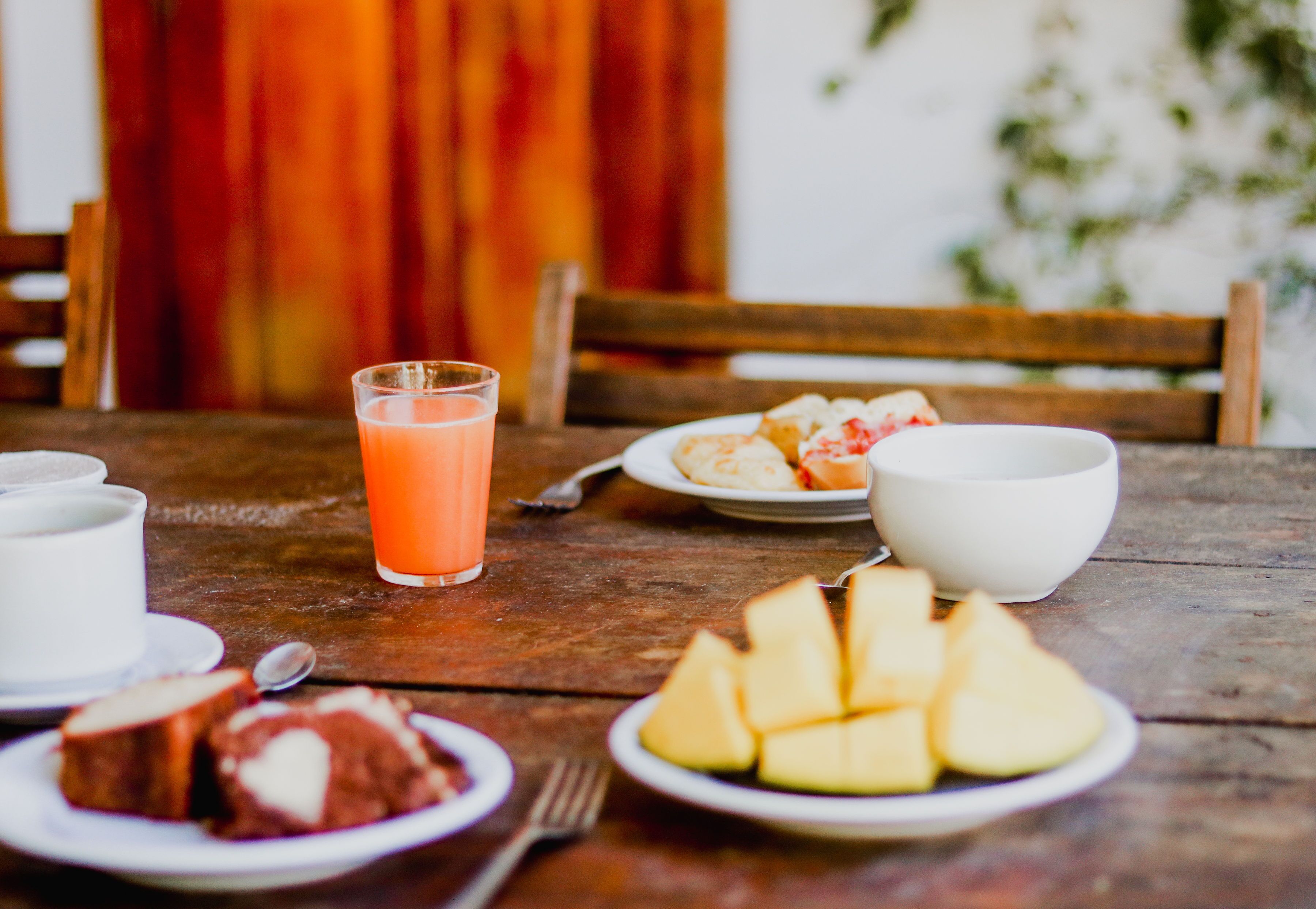 A wooden table with breakfast items including a glass of orange juice, a plate of fruit, a bowl, and a slice of cake.
