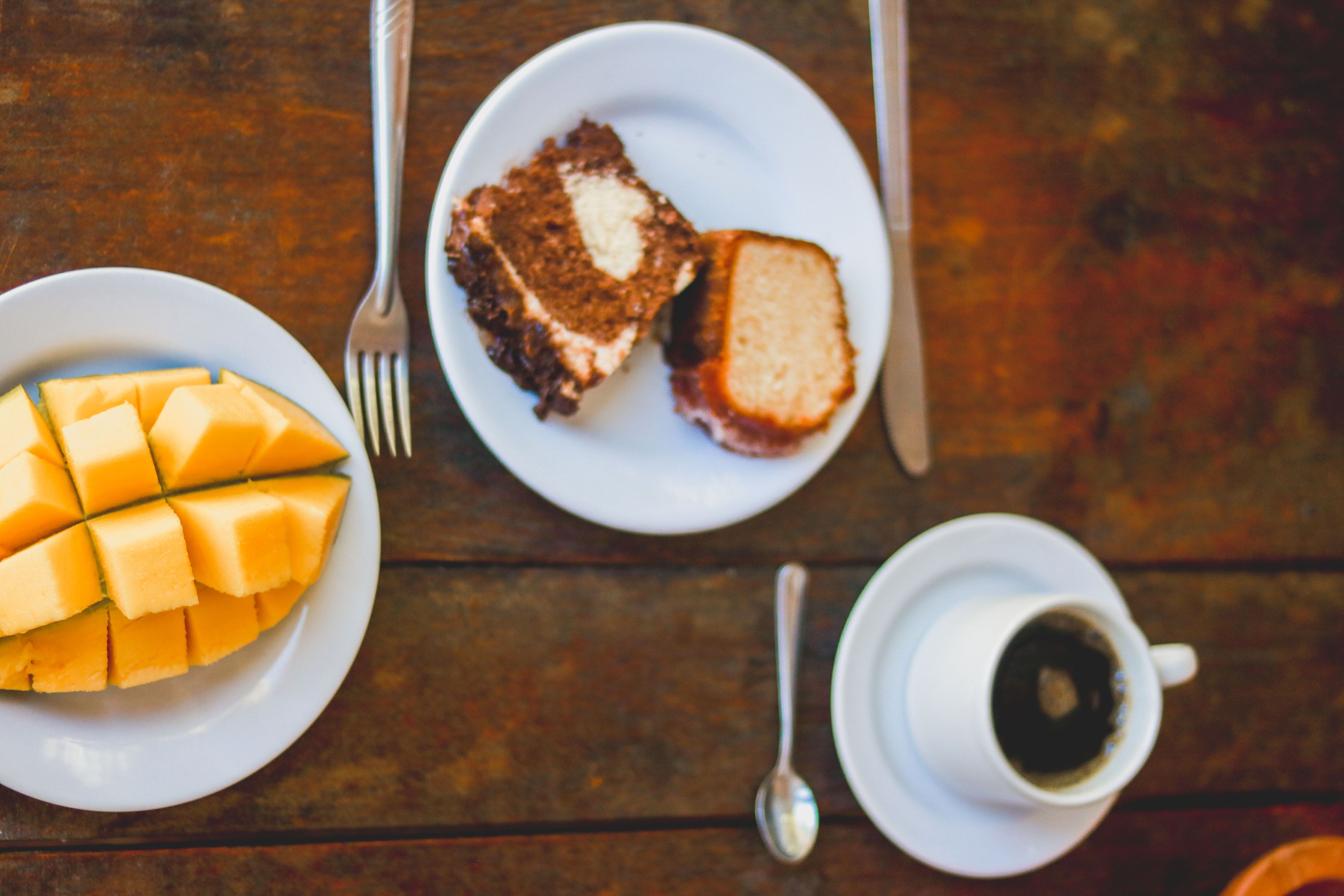A table with a plate of mango slices, a slice of cake, a cup of coffee, and cutlery.