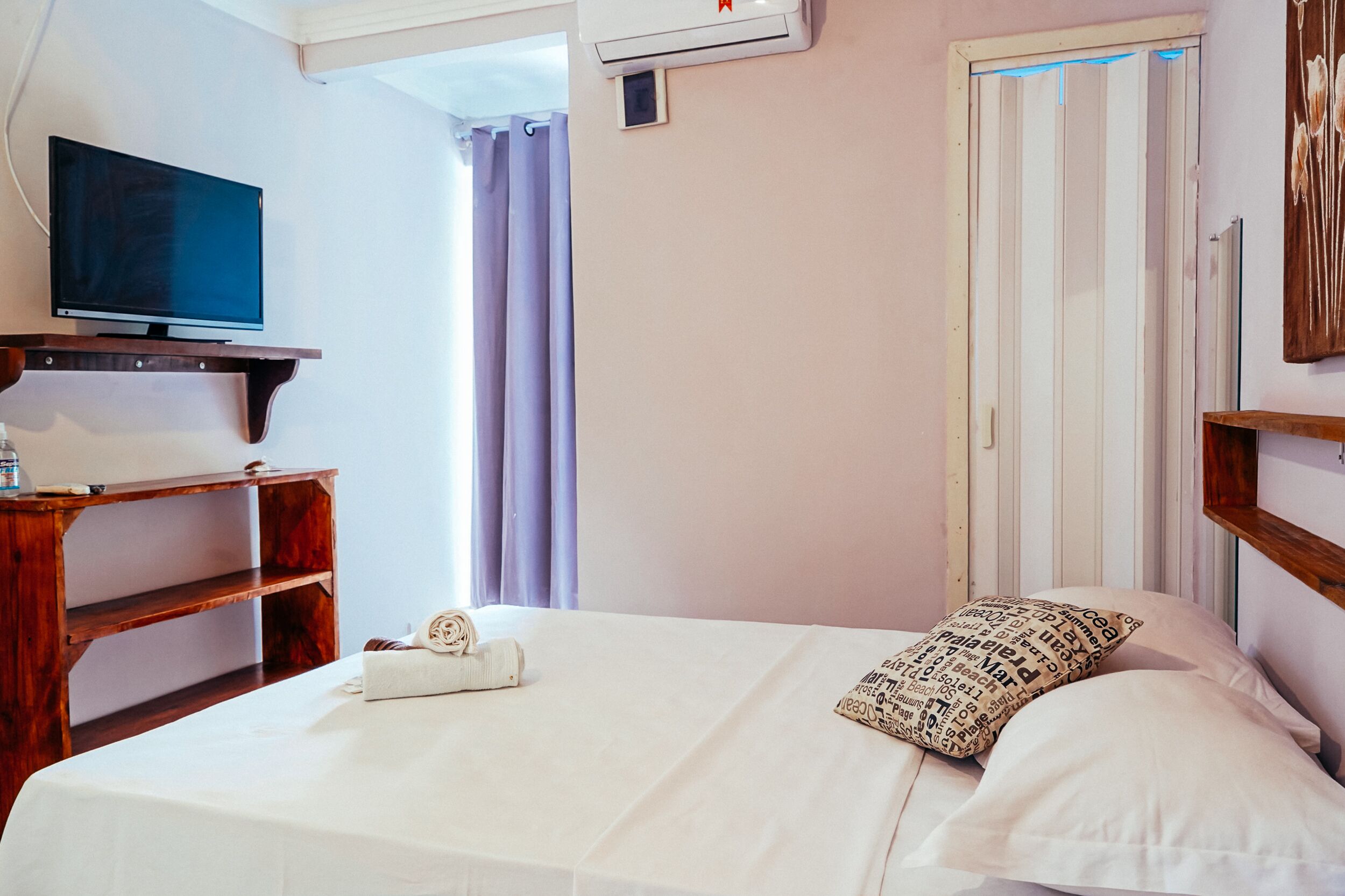 A hotel room with a bed, a TV on a wooden shelf, a window with curtains, and an air conditioning unit.