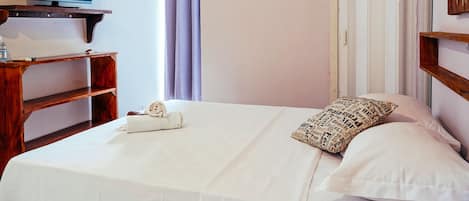 A hotel room with a bed, a TV on a wooden shelf, a window with curtains, and an air conditioning unit.