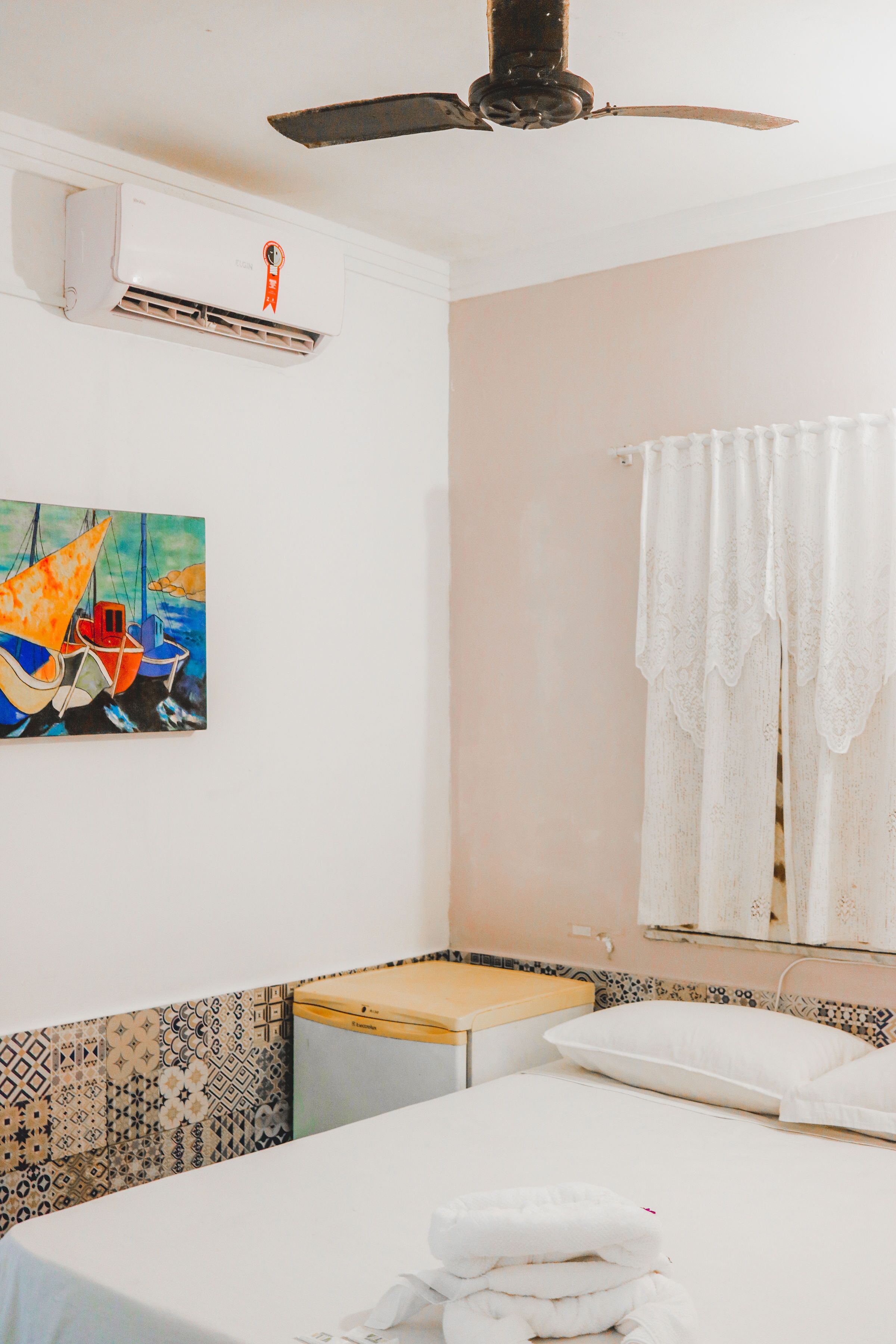 A hotel room with a bed, a ceiling fan, an air conditioning unit, a painting of boats, a window with a white curtain, and a tiled backsplash.