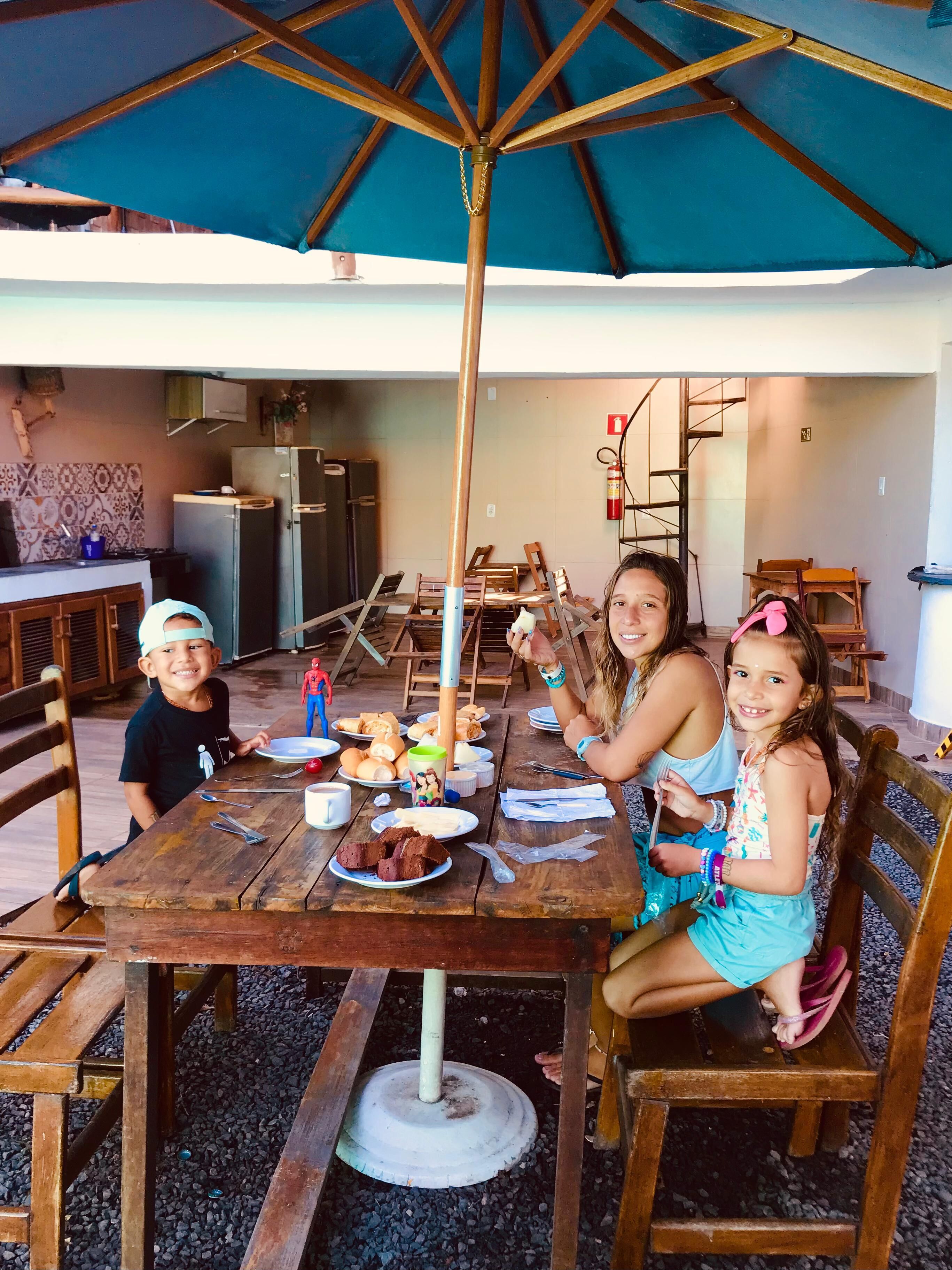 An outdoor dining area with wooden tables and chairs, a blue umbrella, and a kitchen in the background.