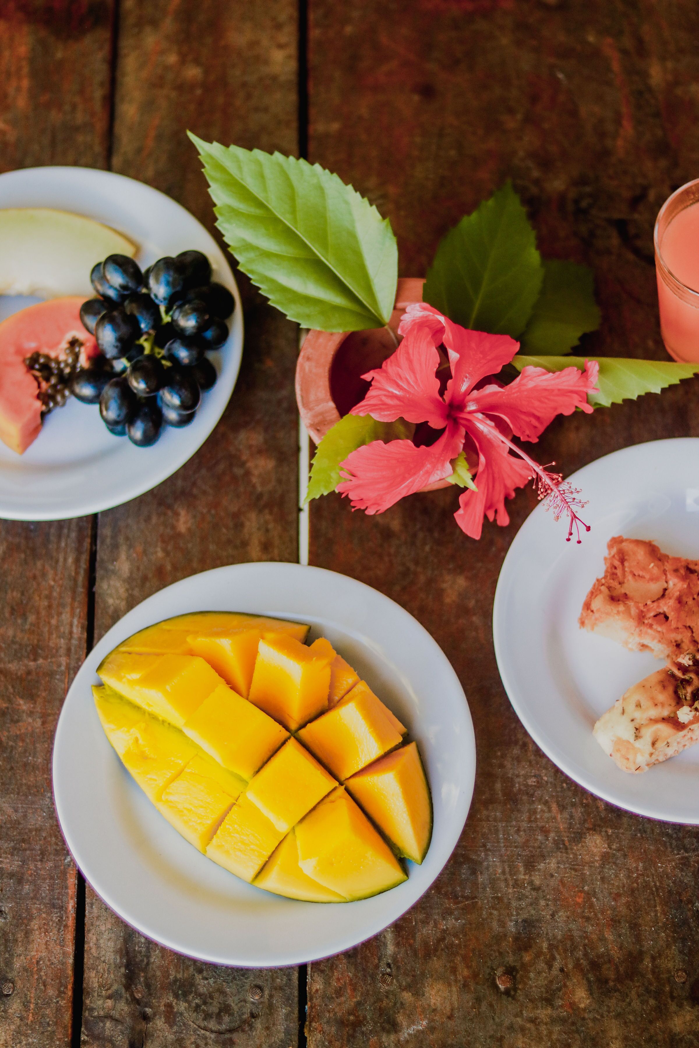 A wooden table with plates of sliced mango, grapes, and a pink drink.