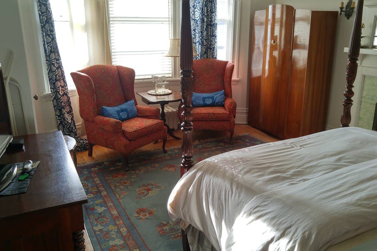 A hotel room with a bed, two armchairs, a small table, a desk with a computer, and a fireplace.