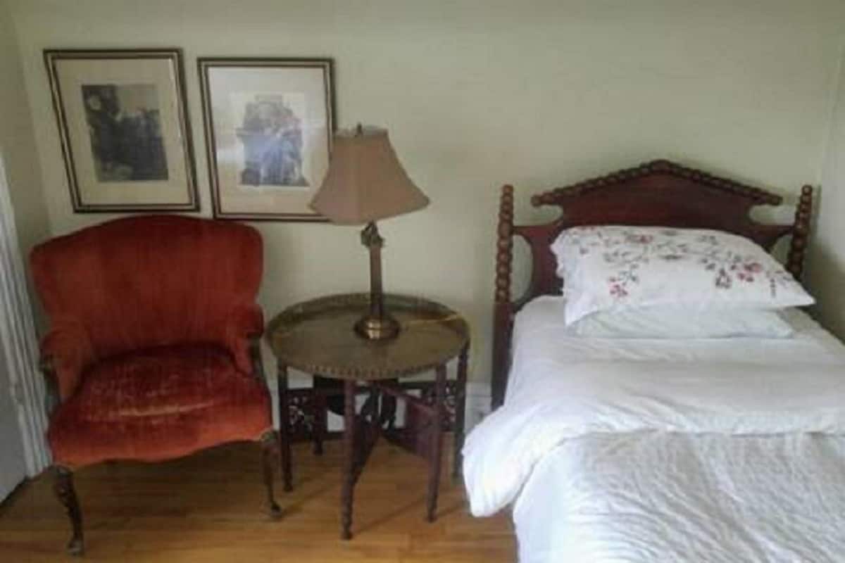 A room with a wooden bed, a red armchair, a small round table with a lamp, and two framed pictures on the wall.