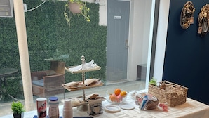 A table set with food and drinks, a wicker basket, and a window with a view of a green hedge and a door. - Dunns Josephine Hotel (Miami)