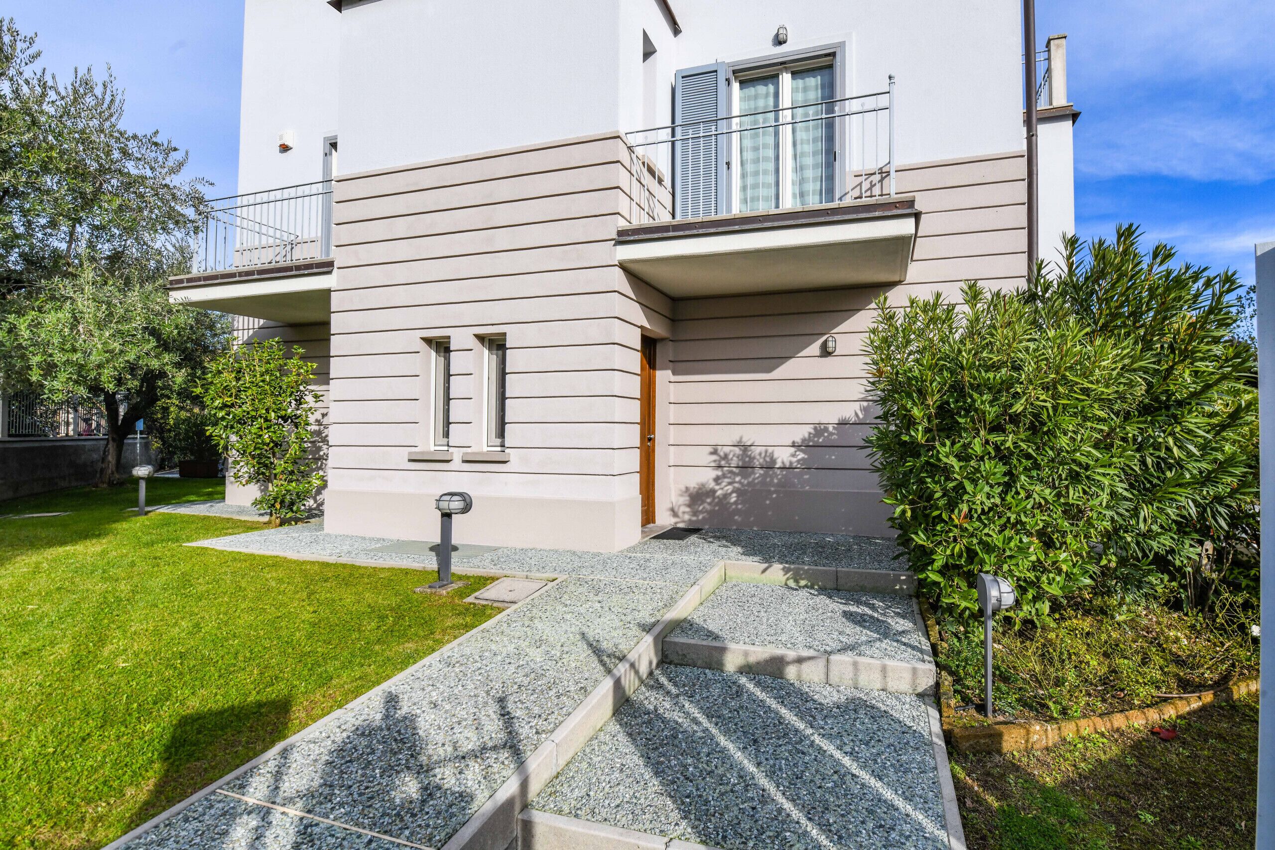 A modern two-story residential building with a flat roof, a balcony with metal railings, and a small front garden with a gravel pathway leading to the entrance.