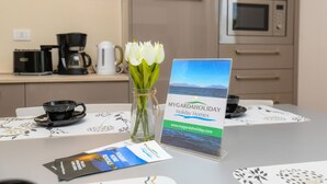 Un coin cuisine équipé d’une machine à café, d’une bouilloire et d’un vase avec des fleurs blanches, le tout posé sur une table. Sur la table se trouve également une brochure intitulée « MYGARDAHOLIDAY, locations de vacances ».