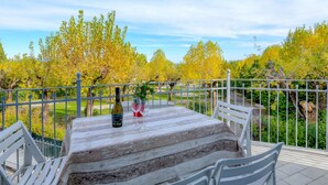 Une table avec une bouteille et des verres, entourée de chaises blanches, donnant sur un jardin avec des arbres et un sentier.