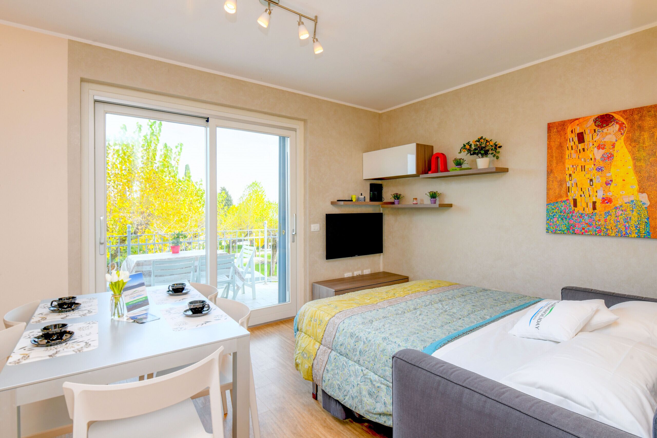 A modern hotel room with a bed, a desk with a chair, a TV, and a balcony with a table and chairs.