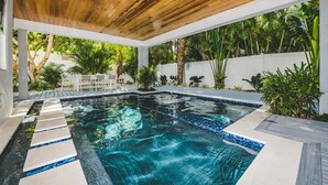 A swimming pool with clear blue water, surrounded by lush greenery and a wooden ceiling. - Luxury Coastal Estate with Rooftop Deck, Pool and Gulf Views (Bradenton Beach)