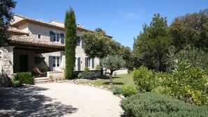A stone house with a tiled roof, a gravel driveway, and a well-maintained garden with various shrubs and trees. - Stone Provencal farmhouse with panoramic view & swimming pool- Saint-Restitut (Saint-Restitut)
