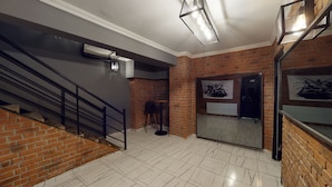 Una escalera con barandilla metálica, una silla negra, una mesita y un espejo que refleja una puerta y un cartel en una pared de ladrillo.