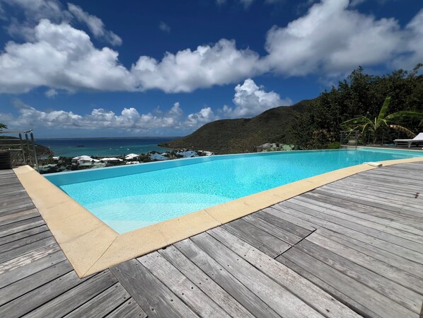A swimming pool with a wooden deck, surrounded by lush greenery and a view of the ocean.