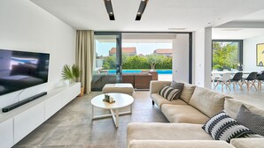 A modern living room with a flat-screen TV, a white coffee table, a beige sofa, and a glass door leading to a pool area. - Villa Lodi - new modern villa  few steps from beach with heated pool (Kaštel Štafilić)