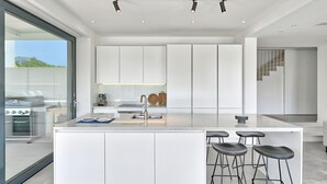 A modern kitchen with white cabinetry, a central island with stools, and a view of the outdoors. - Villa Lodi - new modern villa  few steps from beach with heated pool (Kaštel Štafilić)