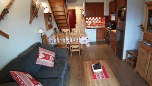 Ein kompakter Wohnraum mit Sofa, Esstisch, Stühlen, einer Küchenzeile mit Holzausbau und Geräten sowie einer Treppe.