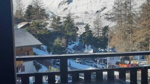 Blick aus einem Fenster auf eine verschneite Berglandschaft mit einer Holzterrasse und einem Tisch im Vordergrund.