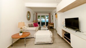Ein modernes Wohnzimmer mit Sofa, Couchtisch und Fernseher. Durch eine Glastür ist ein Blick ins Freie möglich.