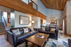 A spacious living area with a large wooden ceiling, a black leather sofa set, a wooden coffee table, and a modern chandelier.