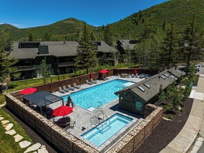 A swimming pool with lounge chairs and umbrellas, surrounded by a stone wall and a mountain backdrop.