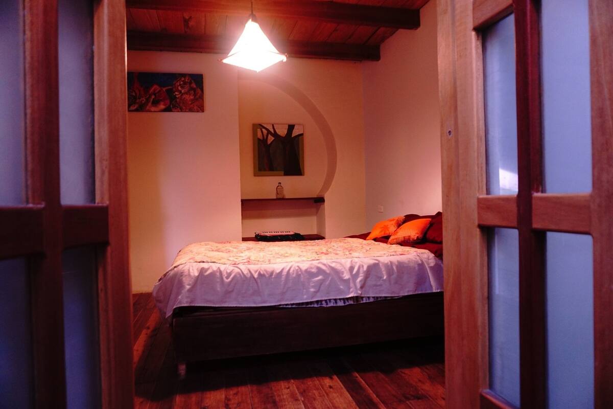 A bedroom with a wooden bed, a hanging light fixture, and a wall-mounted shelf.