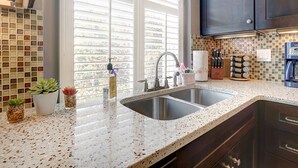 A kitchen with a double sink, dark cabinets, a backsplash with mosaic tiles, and a window with blinds. - Heated Pool Access | 5*Clean | Flex Cancelation (Savannah)
