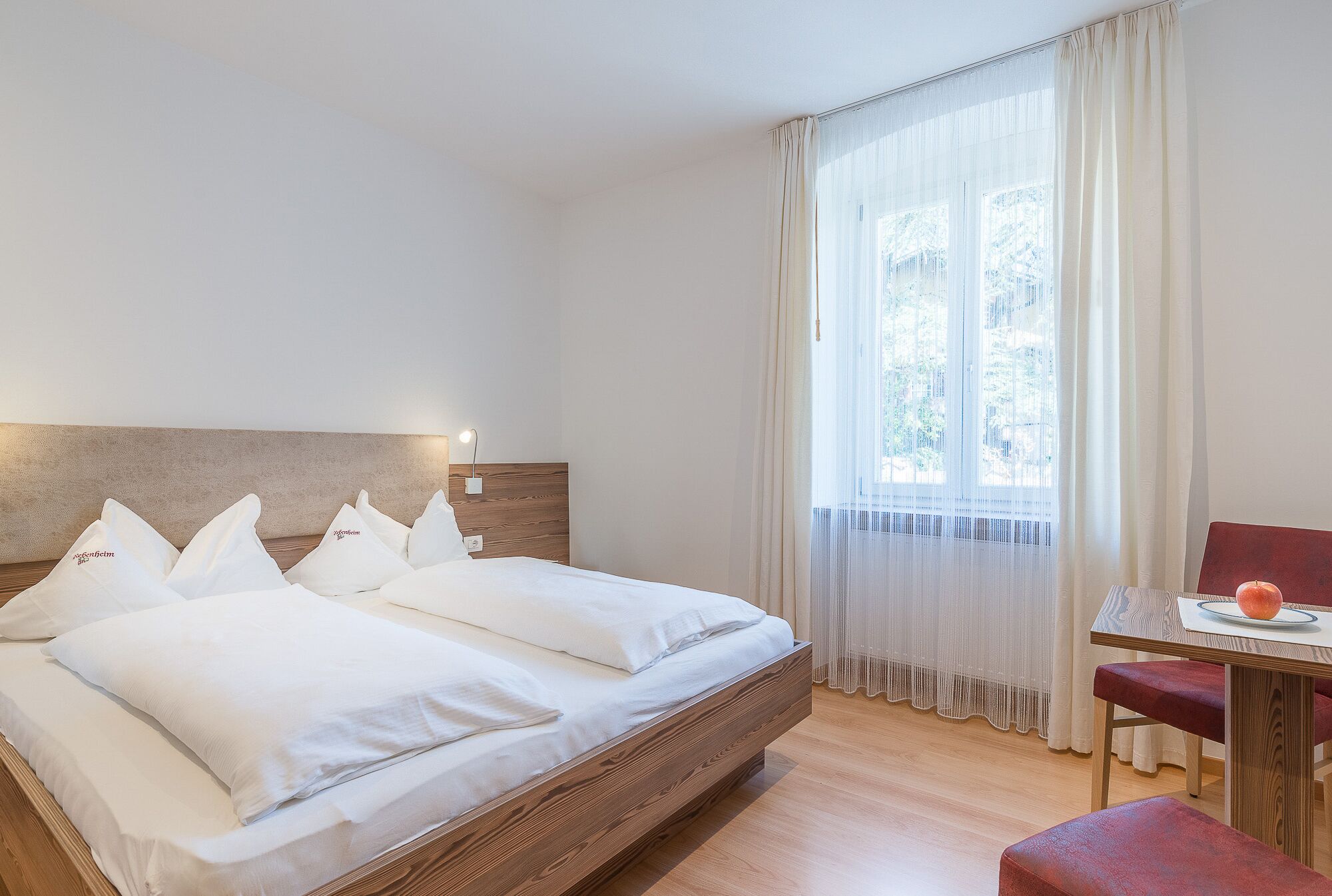 Ein modernes Hotelzimmer mit einem großen Bett, einem Holzkopfende, einem Esstisch mit Stuhl und Blick ins Freie durch leichte Vorhänge.