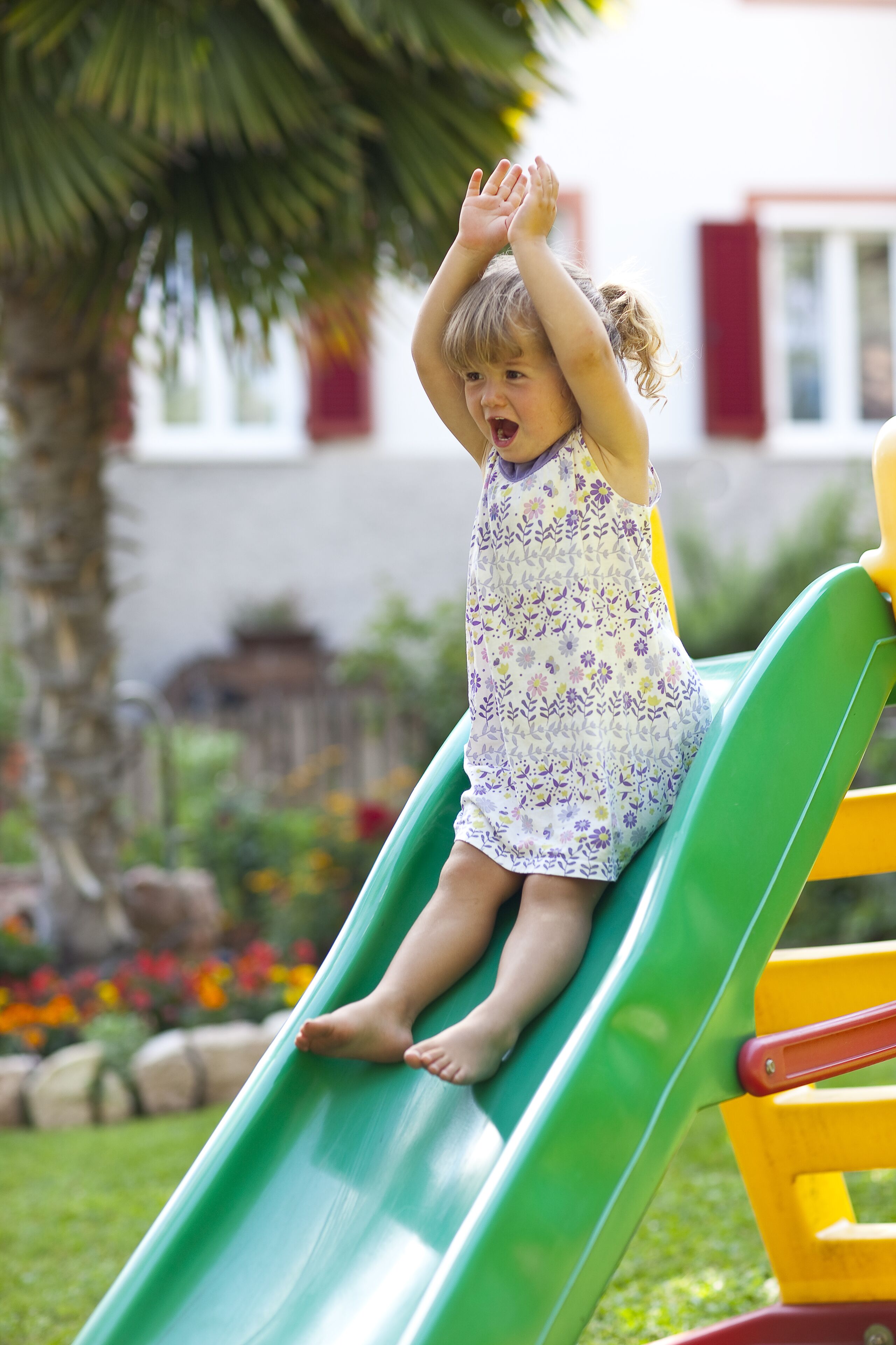 Un enfant sur un toboggan vert dans un terrain de jeux, avec un palmier et des fleurs colorées en arrière-plan.