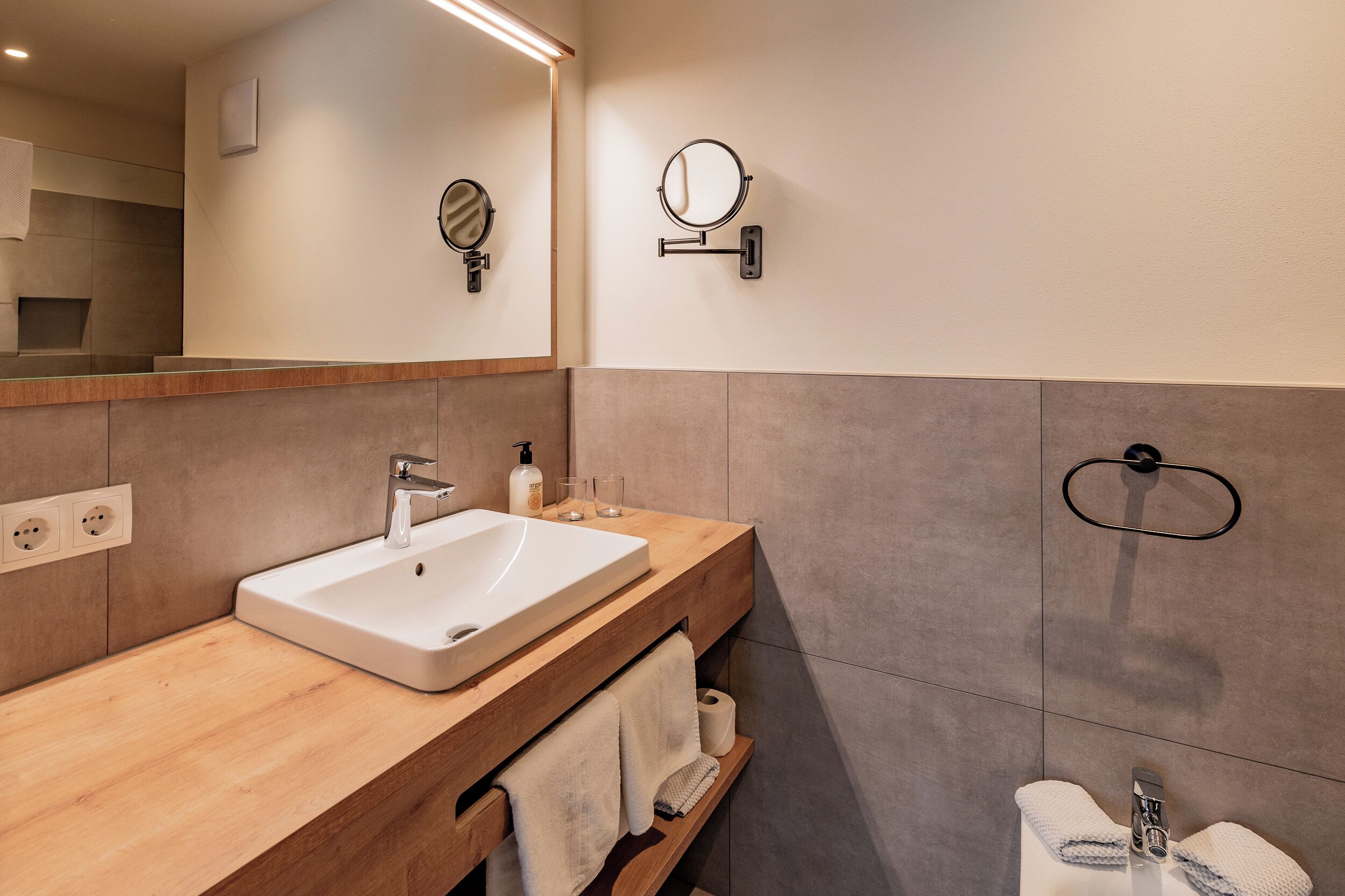 A modern bathroom with a wooden vanity, a large mirror, a wall-mounted towel ring, and a built-in shelf with towels.
