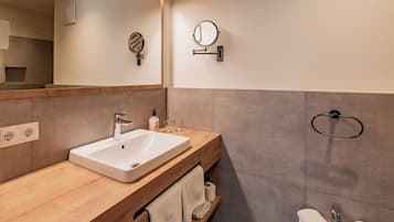 A modern bathroom with a wooden vanity, a large mirror, a wall-mounted towel ring, and a built-in shelf with towels.